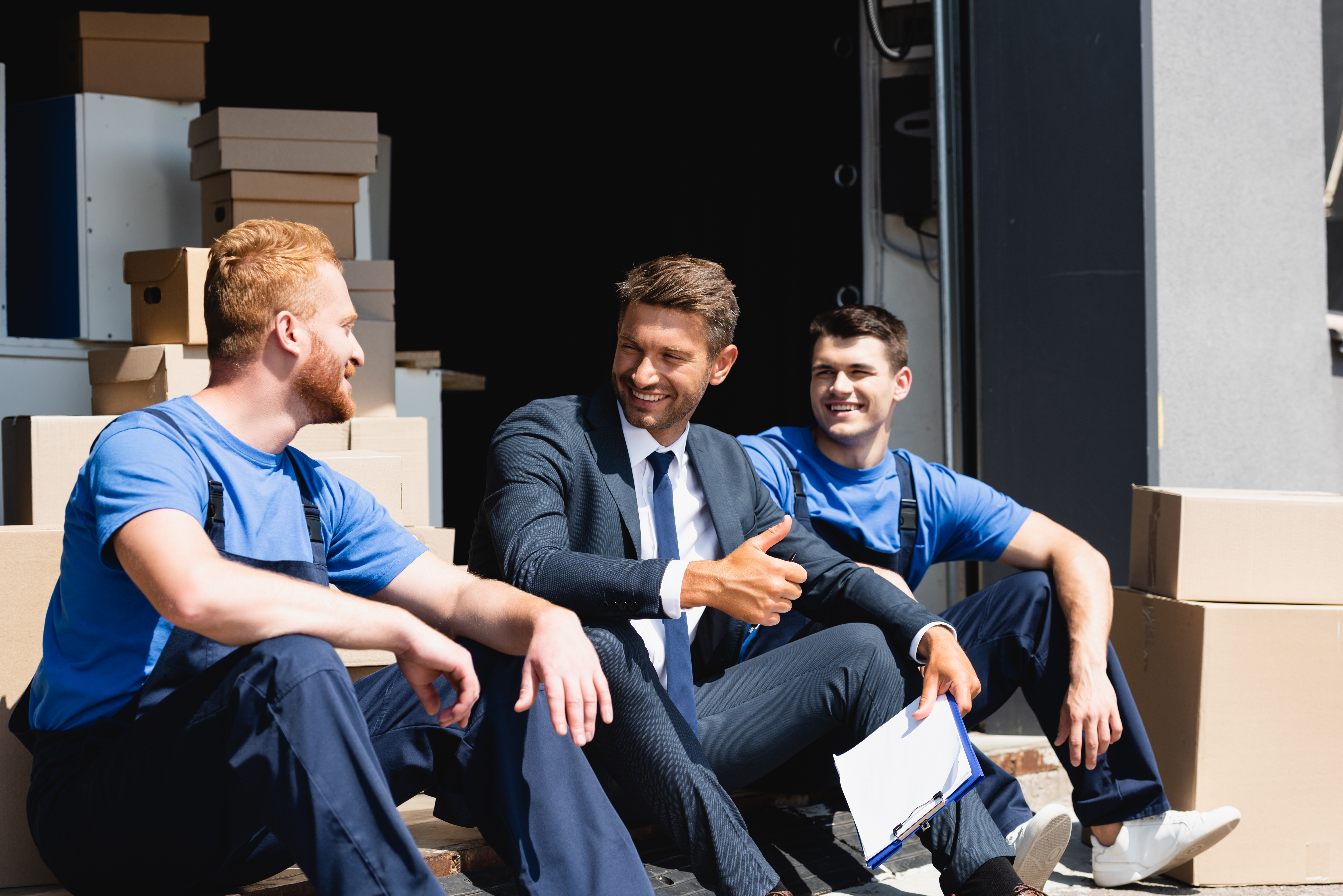 Businessman with clipboard showing like gesture near movers and cardboard boxes in warehouse